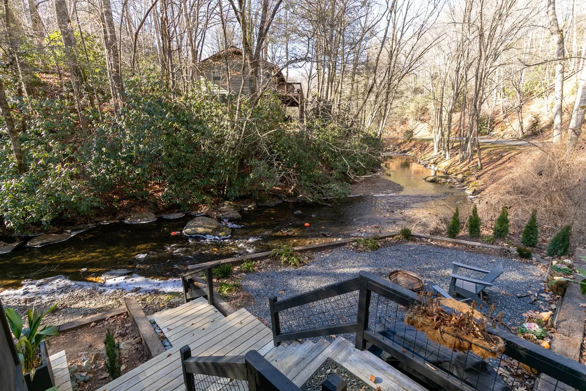 A picturesque view of a meandering creek bordered by verdant greenery and rock formations, with a wooden deck extending close to the water’s edge. The scene is calm and inviting, ideal for fishing, relaxing, or observing wildlife in a natural riverside setting.