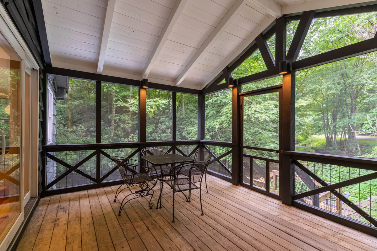 The screened porch features a metal table and chairs set against a backdrop of trees and greenery, offering a comfortable spot for relaxing after a day outside. The porch overlooks surrounding nature, blending indoor comfort with outdoor enjoyment.