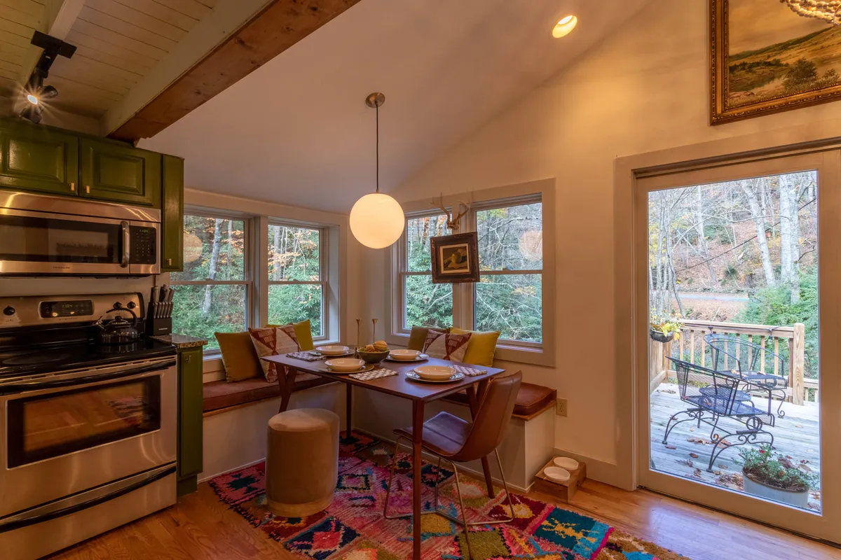 A bright dining area with a large wooden table set for a meal, green kitchen cabinets, and expansive windows framing a lush river view. Decorative pendant lighting adds a welcoming touch, and the natural scenery just beyond the glass emphasizes the riverside setting.