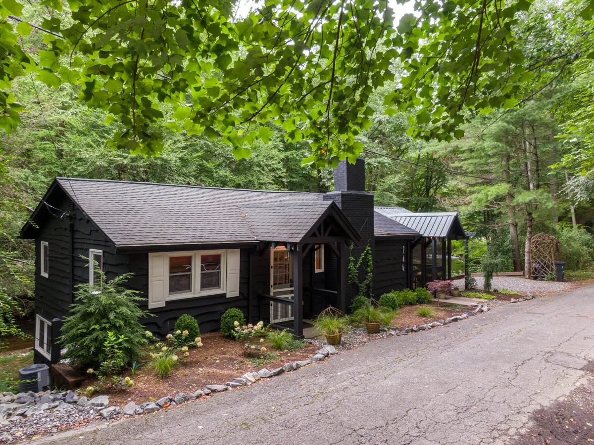 A calm exterior view of the cottage's black wooden façade is surrounded by greenery, with a stone pathway leading to a cozy front porch. The natural setting highlights the tranquility and inviting atmosphere of the property, suggesting peaceful mornings or evenings spent outdoors.