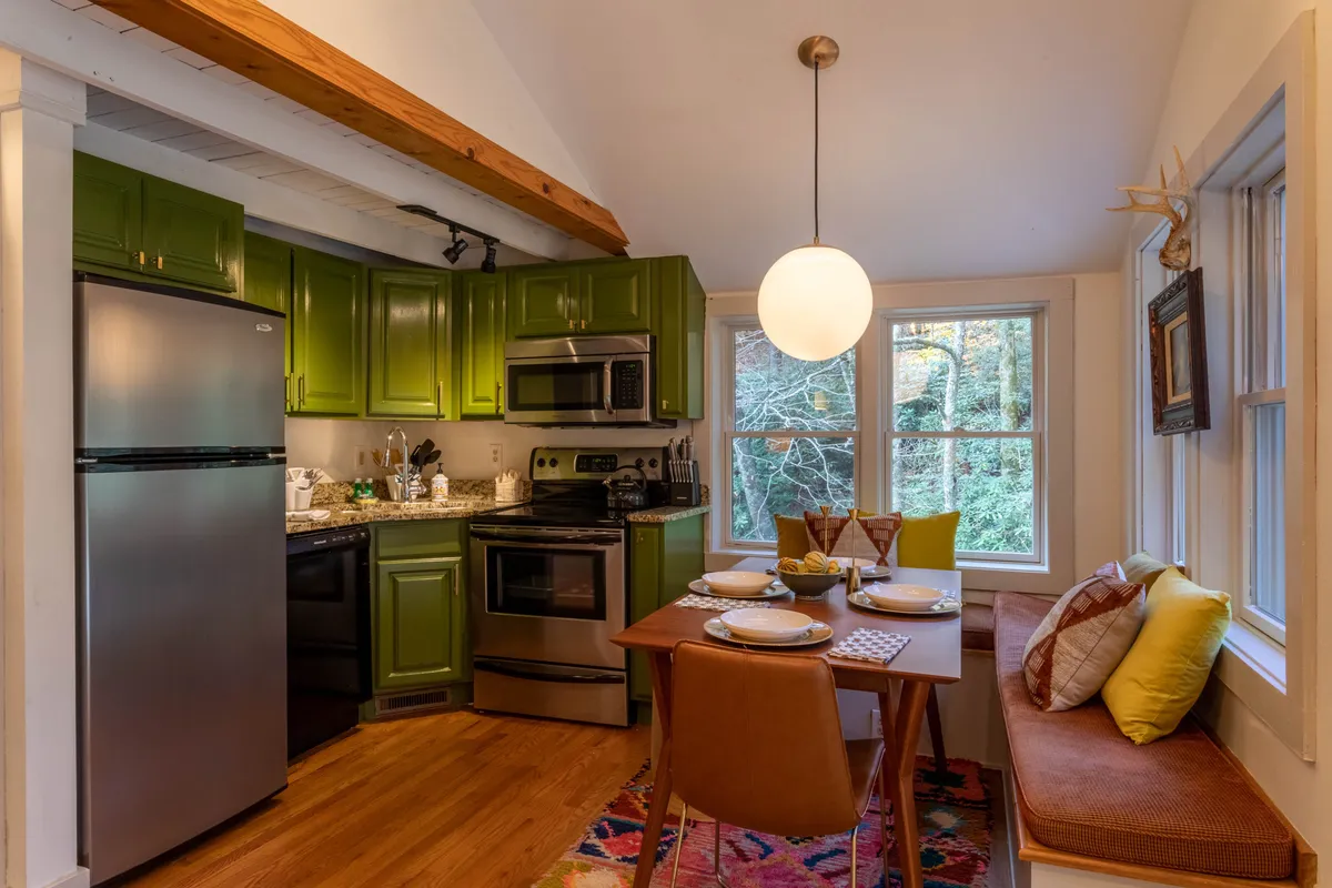 This image features a kitchen and dining area with green cabinets, a spacious dining table, window seating, and a warm pendant light overhead. The arrangement suggests a space that is both practical for meal preparation and inviting for gatherings, with natural light streaming in.