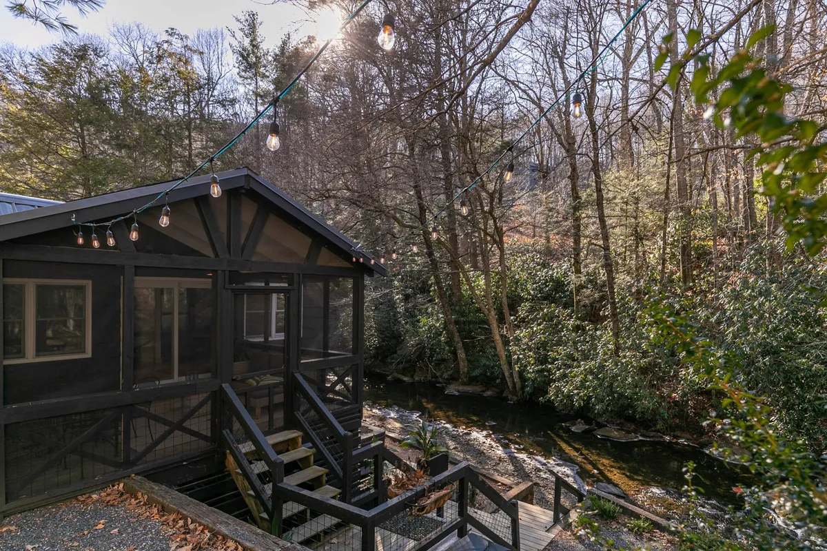 A serene view of the screened porch illuminated with string lights, overlooking a leafy backdrop and a gently flowing creek. The inviting space suggests relaxing evenings under the stars, with the sounds of nature close by.