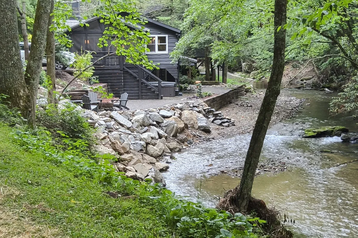 The image shows a charming, updated cottage nestled by the edge of the Middle Fork of the New River. The cottage features a deck overlooking the gently flowing stream, with comfortable seating perfect for morning coffee or evening relaxation. The tranquil setting is enhanced by natural surroundings—lush greenery, sunset-lit skies, and the promise of waterside activities. A fire pit area invites guests to gather in the evenings, making the space ideal for unwinding after a day of outdoor adventures.
