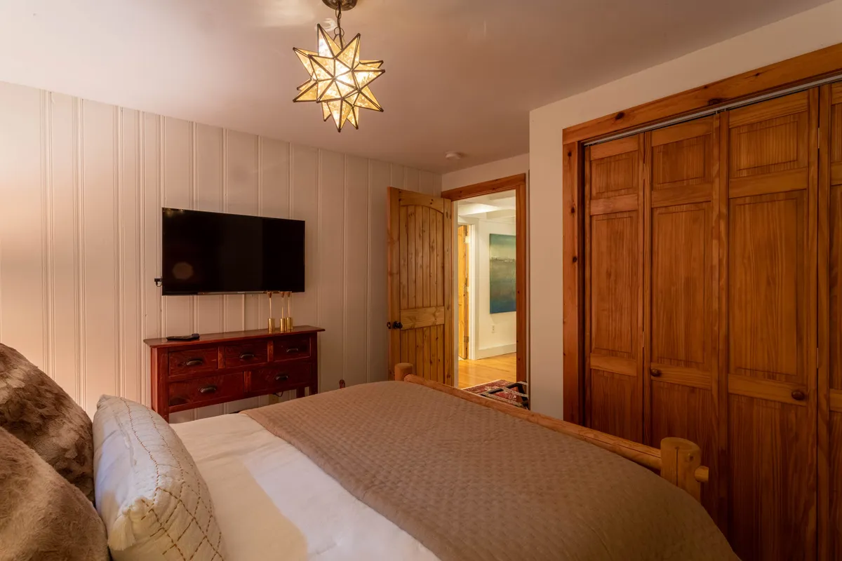 A tastefully decorated bedroom with a textured blanket on the queen bed, a wooden dresser, a wall-mounted TV, and a distinctive wooden star pendant light, creating a cozy and intimate atmosphere.