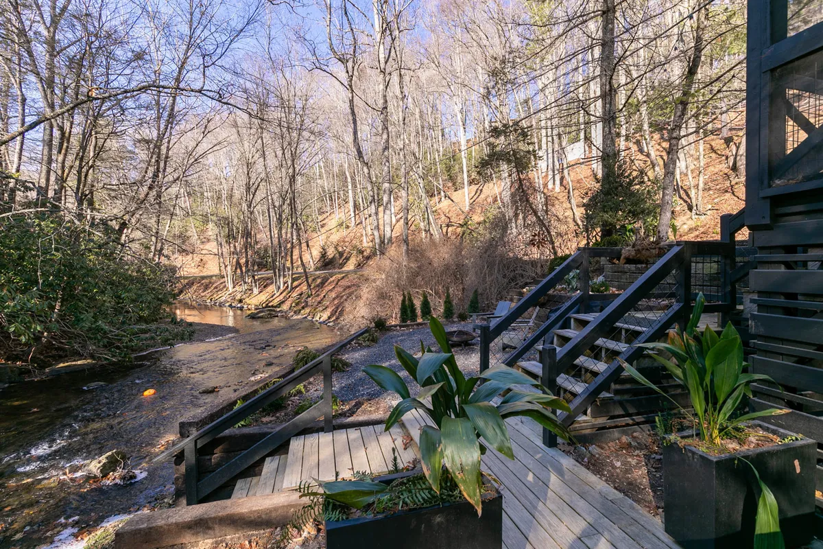 A tranquil view of wooden stairs leading down to a stream, framed by leafy trees and potted plants. The image hints at private, easy access to natural areas right from the property, inviting relaxing strolls and moments by the water.