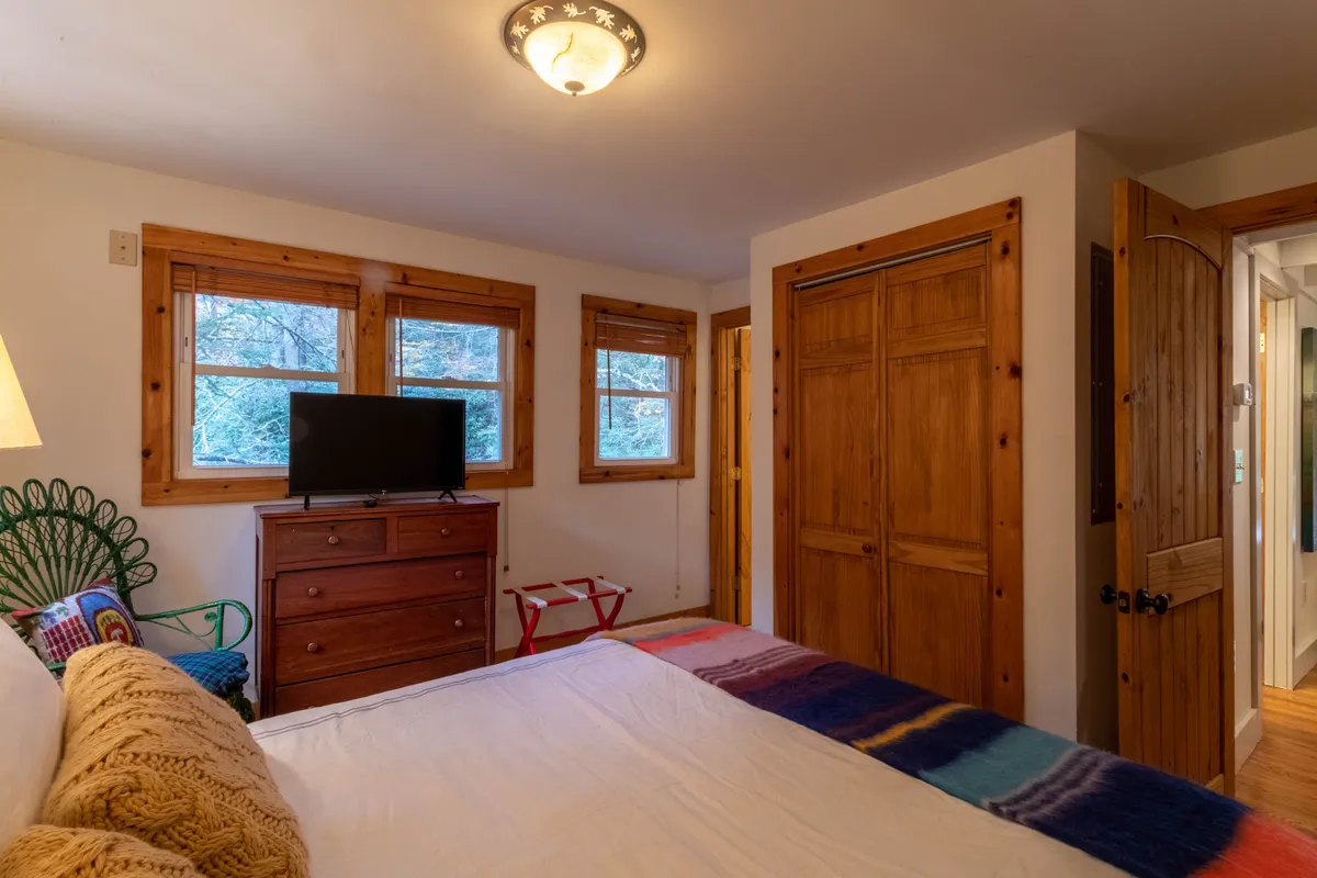A warm, inviting bedroom featuring a queen bed adorned with a colorful blanket, a wooden dresser, a TV, a green chair, and large windows letting in abundant natural light. The overall setting exudes a serene, restful vibe, suggestive of a peaceful retreat.