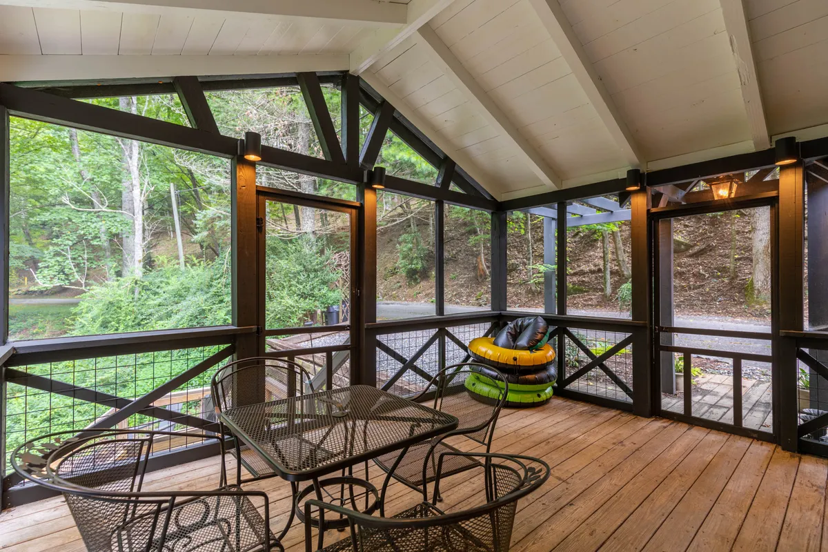 The image features a screened-in porch furnished with a wooden table and chairs, inviting guests to relax and enjoy meals or conversations. A colorful inner tube hints at nearby water activities, while the porch looks out onto lush greenery, seamlessly blending indoor comfort with an outdoor riverside environment.