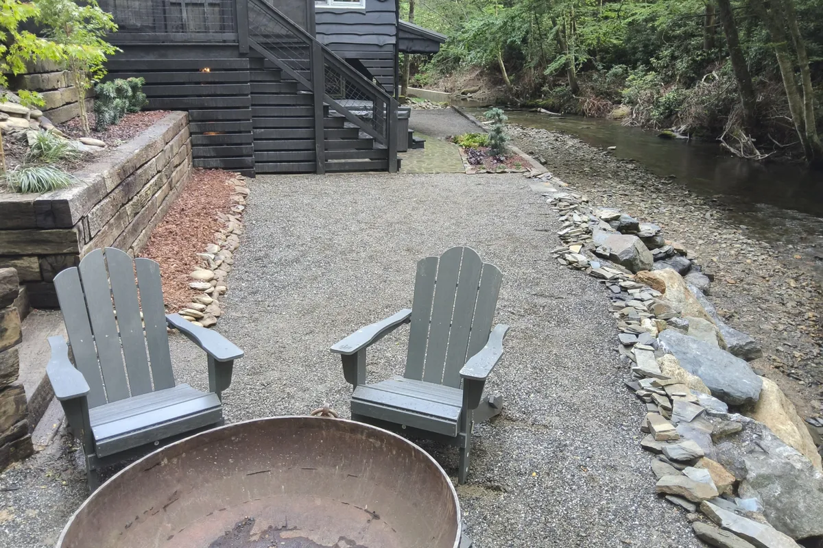 This image captures two gray Adirondack chairs positioned around a fire pit on a stone pathway, set along the riverbank. The view includes the gentle river, lush greenery, and an inviting outdoor seating area ideal for relaxing or storytelling by the fire.
