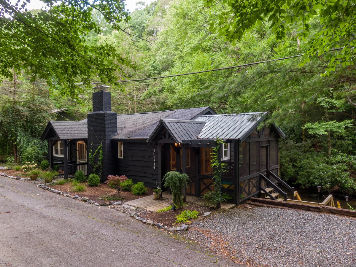 A welcoming exterior view of Anglers Cottage showcasing its black wooden façade, stone chimney, and a screened porch nestled amid lush greenery. The setting is private and enveloped by nature, hinting at the tranquility and seclusion the cottage provides for couples and small groups.
